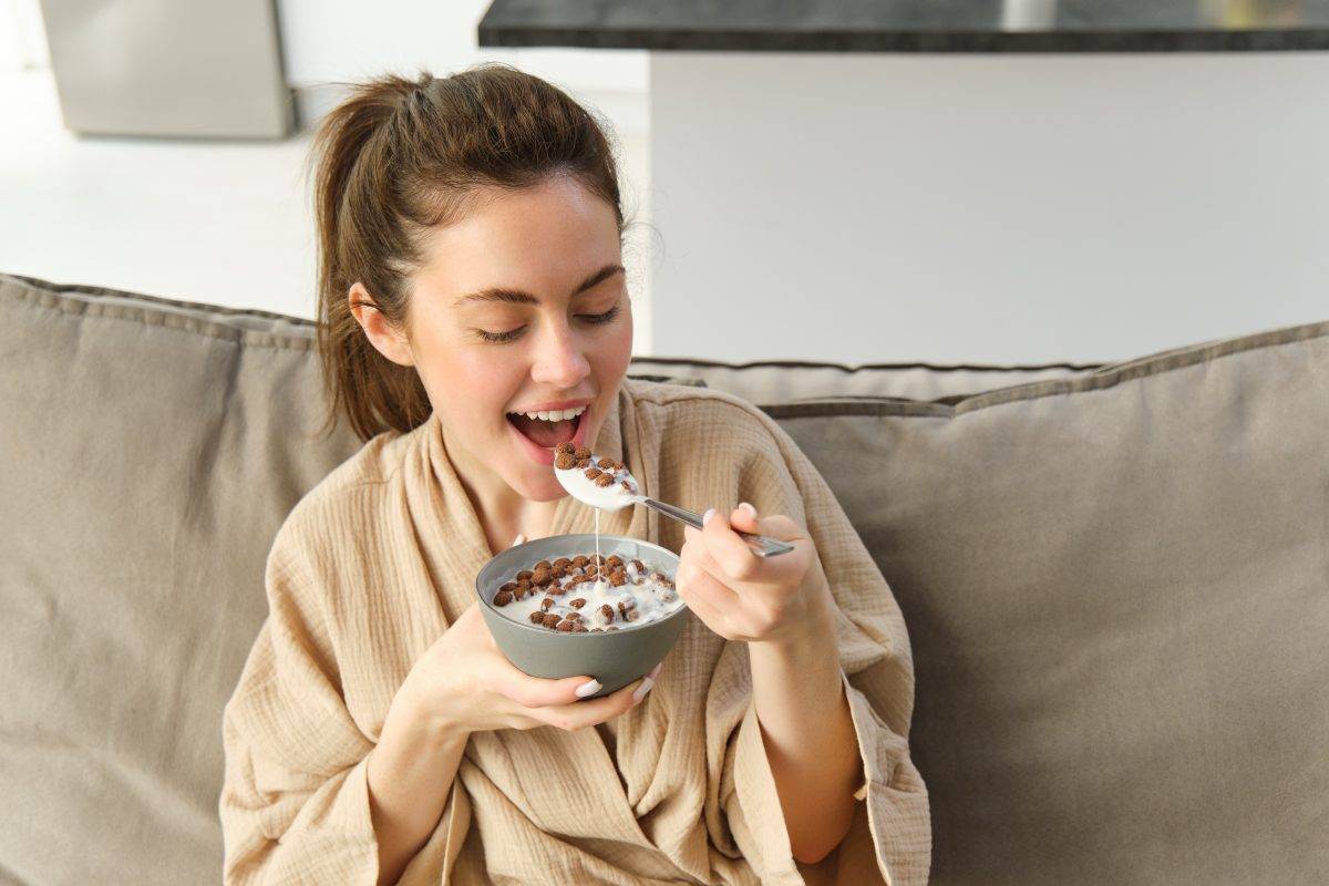 Portrait of gorgeous brunette woman sits on sofa, having her breakfast at home, smiling while eating cereals, holding bowl and spoon and laughing.